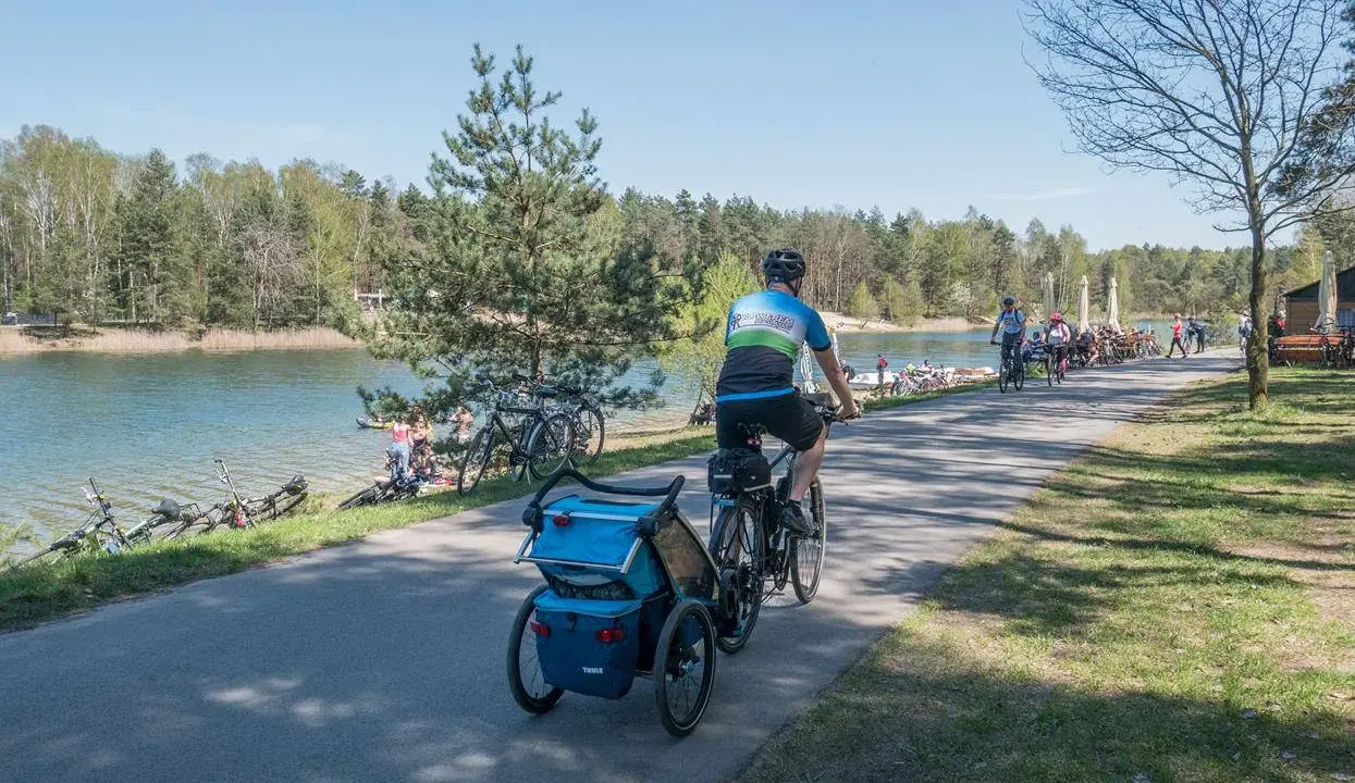 Rodzina na rowerach z przyczepką nad jeziorem Świerklaniec. W tle inni rowerzyści i plażowicze.