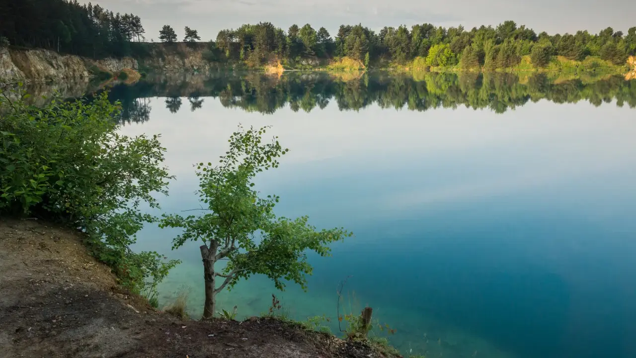 Spokojne jezioro wapienne z odbijającymi się w wodzie drzewami i niebem. Brzeg porośnięty zielenią, z widocznymi skalnymi zboczami.