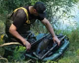Wędkarz w wodoodpornych spodniach i kamizelce z żółtymi szelkami ostrożnie wyciąga karpia z siatki. Obok leżą worki karpiowe.