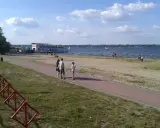 Rodzina spaceruje brzegiem jeziora Balewskiego. Na wodzie żaglówki, na brzegu barka z restauracją.