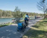 Rodzina na rowerach z przyczepką nad jeziorem Świerklaniec. W tle inni rowerzyści i plażowicze.