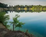 Spokojne jezioro wapienne z odbijającymi się w wodzie drzewami i niebem. Brzeg porośnięty zielenią, z widocznymi skalnymi zboczami.