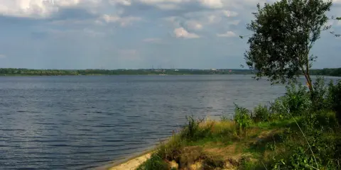 Szerokie jezioro z piaszczystym brzegiem i drzewem w oddali, pod zachmurzonym niebem.