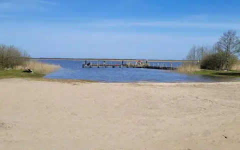 Plaża nad jeziorem Wicko, z pomostem, na którym stoją ludzie. W oddali widać trzciny i drzewa.