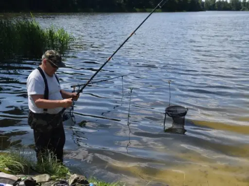 Wędkarz łowi ryby na jeziorze wierzbiczańskim. W wodzie stoi podbierak, a w tle widać zarośla i las.