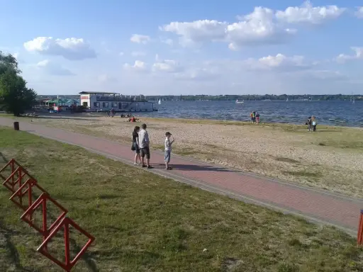 Rodzina spaceruje brzegiem jeziora Balewskiego. Na wodzie żaglówki, na brzegu barka z restauracją.
