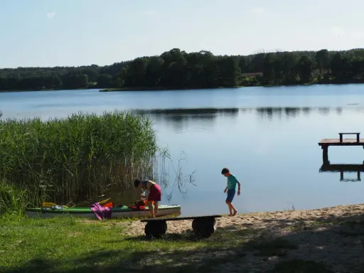 Dzieci szykują kajak nad spokojnym jeziorem w gminie Morąg. W tle las i drewniane molo.