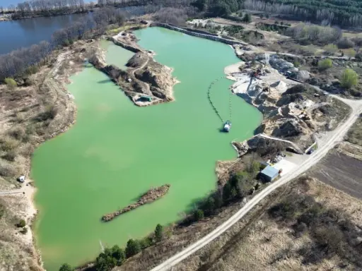 Widok z lotu ptaka na turkusowe jezioro zajączkowskie, otoczone piaszczystymi skarpami i zielenią. Widać sprzęt wydobywczy i łódź.