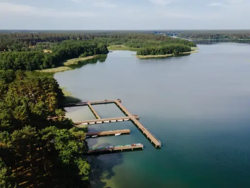 Drewniane pomosty na spokojnym jeziorze Wiartel, otoczonym gęstym lasem. Woda jest przejrzysta, a na pomostach stoją łódki i kajaki.