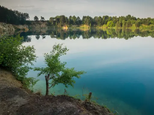 Spokojne jezioro wapienne z odbijającymi się w wodzie drzewami i niebem. Brzeg porośnięty zielenią, z widocznymi skalnymi zboczami.