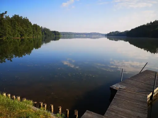 Spokojne jezioro Sulęczyno o poranku. Drewniane molo zaprasza do podziwiania odbijających się w tafli chmur i zielonych lasów.