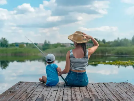 Matka i syn siedzą na drewnianym pomoście, łowiąc ryby. Spokojne łowiska wędkarskie, odbijające się w wodzie chmury.
