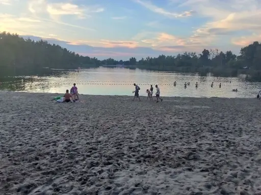 Zachód słońca nad stawem w Jelczu Laskowicach. Ludzie spacerują po piaszczystej plaży i kąpią się w wodzie.