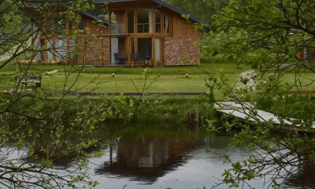 Nowoczesny domek ze stawem rybnym, z drewnianą elewacją i ceglanymi akcentami, odbija się w spokojnej tafli wody.