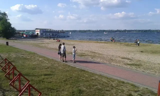 Rodzina spaceruje brzegiem jeziora Balewskiego. Na wodzie żaglówki, na brzegu barka z restauracją.