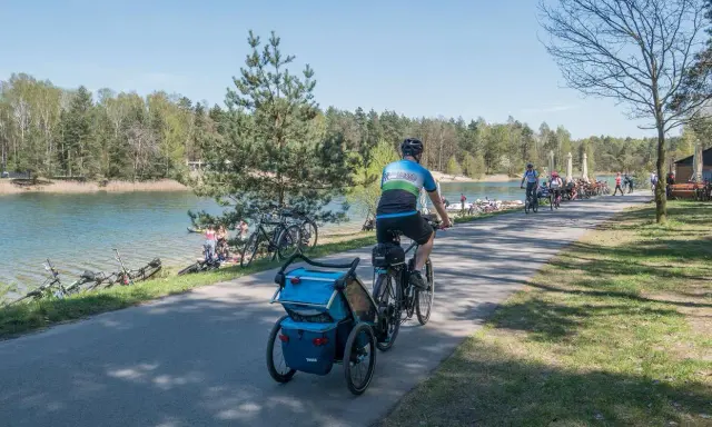 Rodzina na rowerach z przyczepką nad jeziorem Świerklaniec. W tle inni rowerzyści i plażowicze.