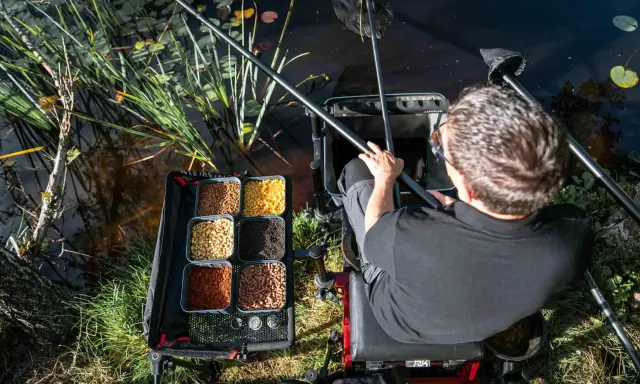 Wędkarz siedzi nad wodą, obok niego tacka z różnymi rodzajami przynęt, w tym gumki do pelletu.