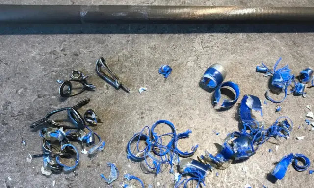 Metalowe przelotki do wędki leżą na szarym blacie, obok niebieskich wiórów i kawałków plastiku.
