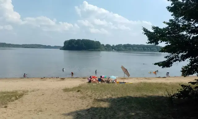 Słoneczny dzień nad jeziorem przeczyckim. Ludzie odpoczywają na piaszczystej plaży, kąpią się i pływają.