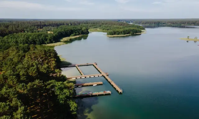 Drewniane pomosty na spokojnym jeziorze Wiartel, otoczonym gęstym lasem. Woda jest przejrzysta, a na pomostach stoją łódki i kajaki.