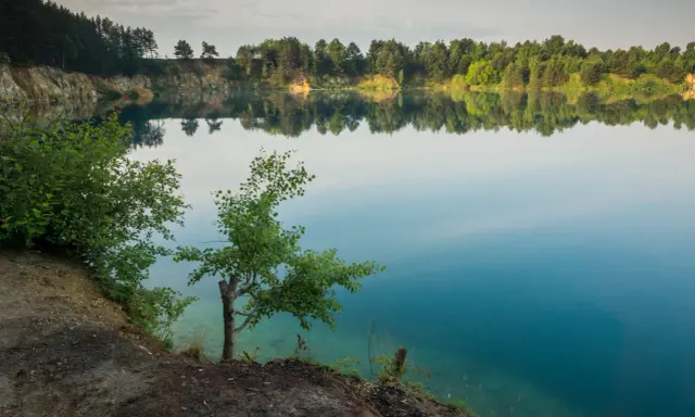 Spokojne jezioro wapienne z odbijającymi się w wodzie drzewami i niebem. Brzeg porośnięty zielenią, z widocznymi skalnymi zboczami.