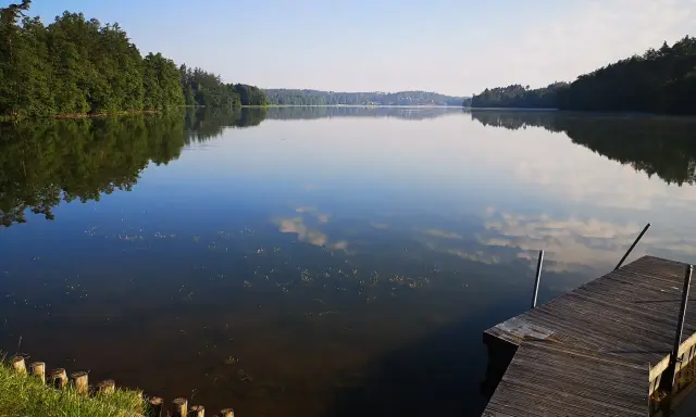 Spokojne jezioro Sulęczyno o poranku. Drewniane molo zaprasza do podziwiania odbijających się w tafli chmur i zielonych lasów.