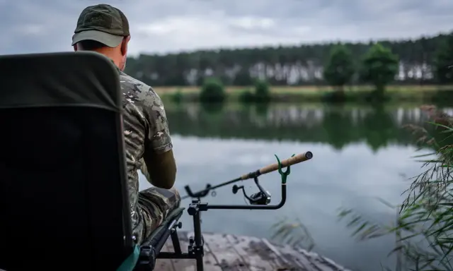 Wędkarz w kamuflażu siedzi na krześle nad jeziorem, gotowy do łowienia. Jego wędka feeder 3m spoczywa na podpórce, czekając na branie.