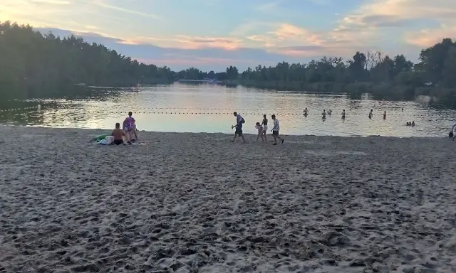 Zachód słońca nad stawem w Jelczu Laskowicach. Ludzie spacerują po piaszczystej plaży i kąpią się w wodzie.
