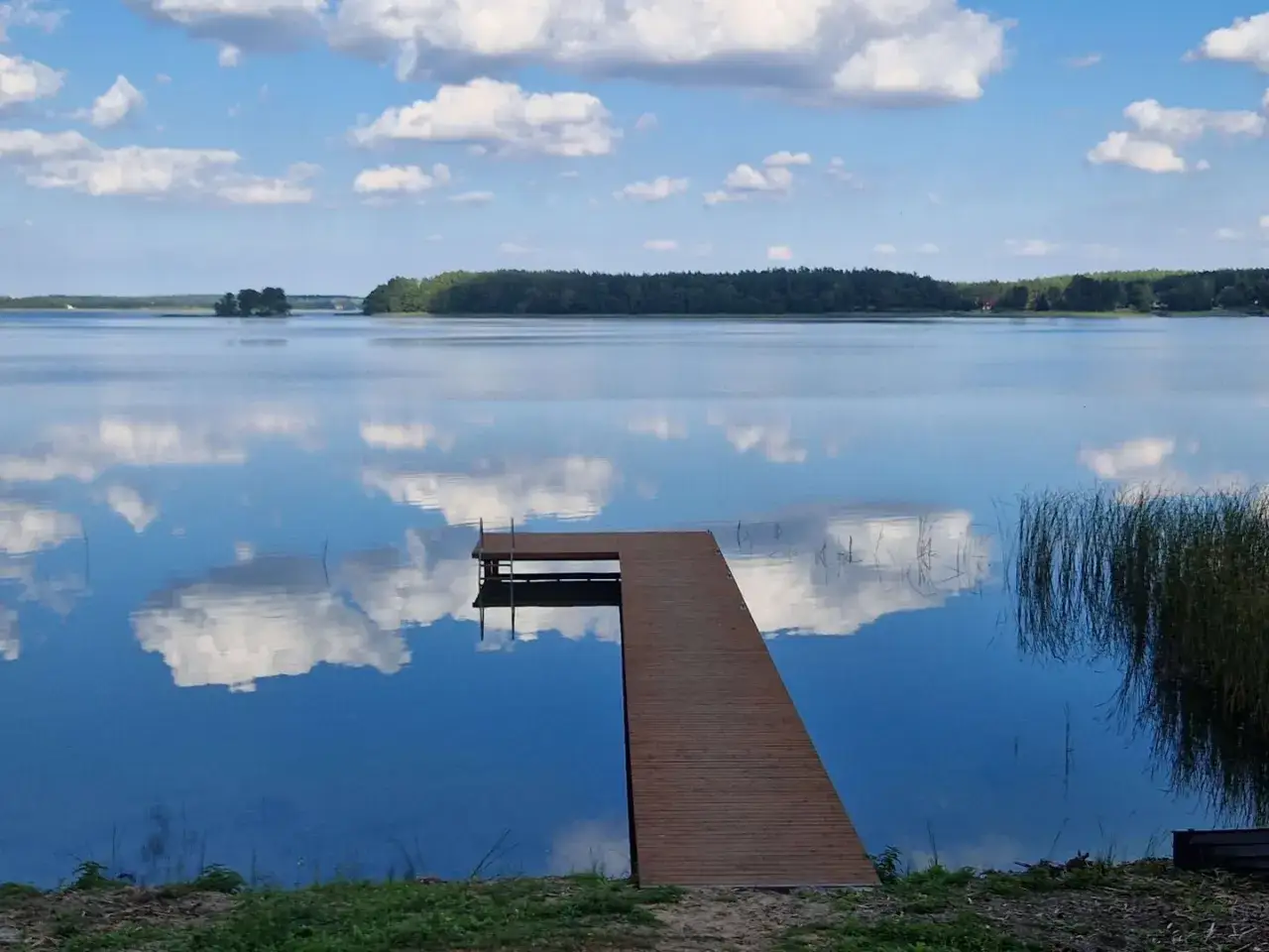 Drewniane molo na spokojnym jeziorze Wilkowskim, odbijające błękitne niebo i białe chmury.
