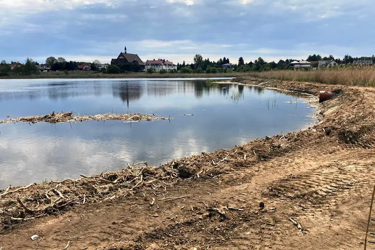 Zalew Zielona Kaleta, spokojna tafla wody odbija chmury. Na brzegu widać ślady prac ziemnych i pozostałości roślinności. W oddali kościół i zabudowania.