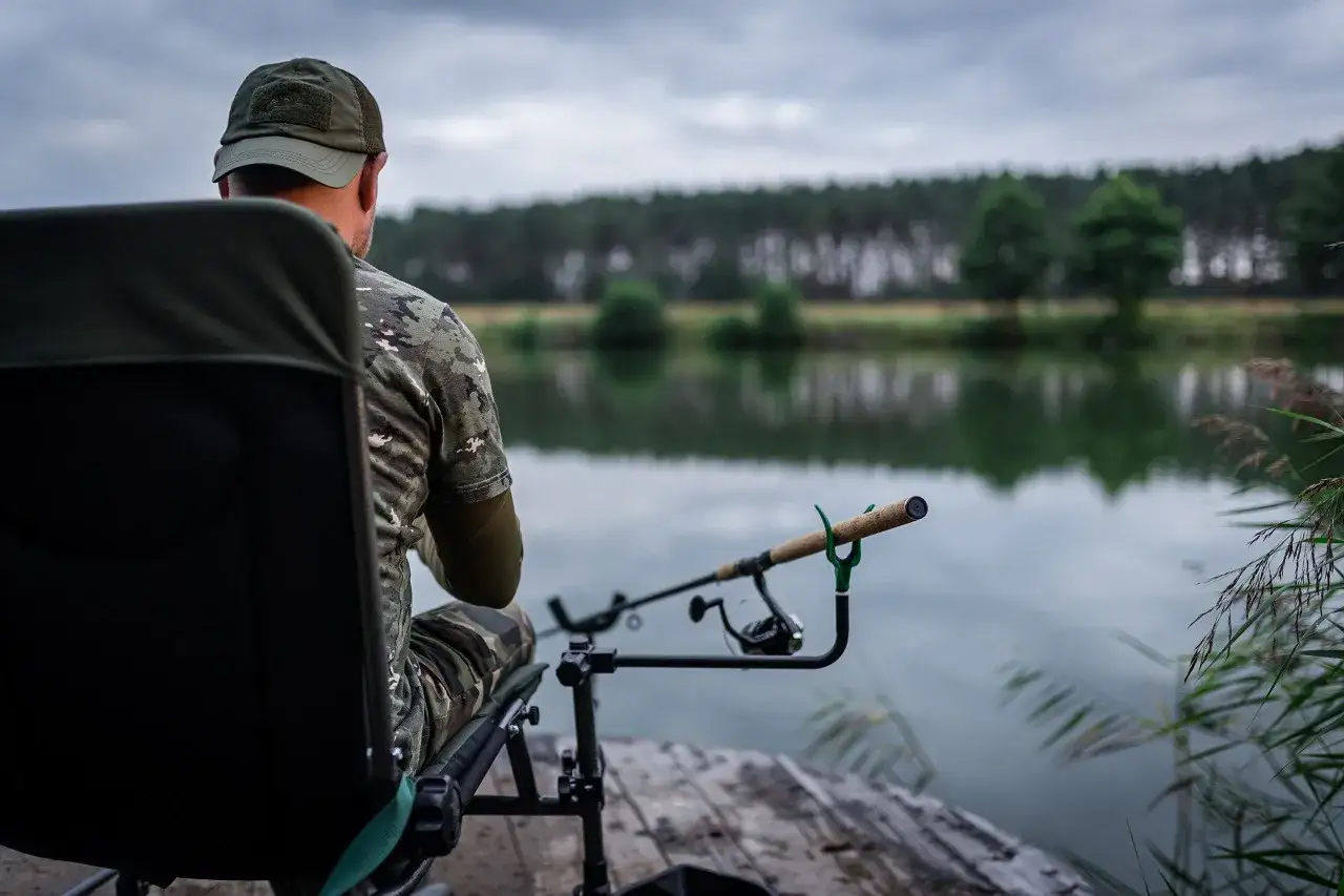 Wędkarz w kamuflażu siedzi na krześle, czekając na branie przy wodzie. Jego wędka feeder spoczywa na podpórce, gotowa do akcji.