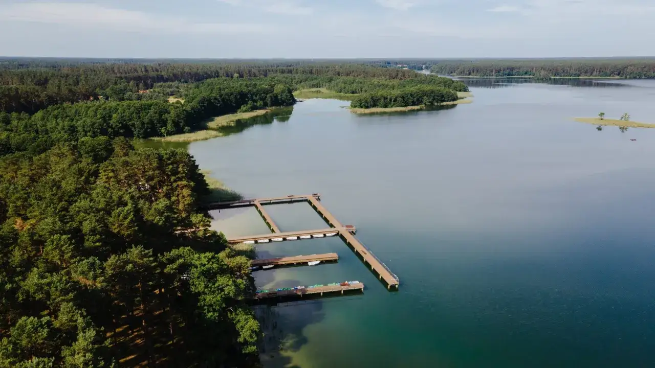 Drewniane pomosty na spokojnym jeziorze Wiartel, otoczonym gęstym lasem. Woda jest przejrzysta, a na pomostach stoją łodzie.