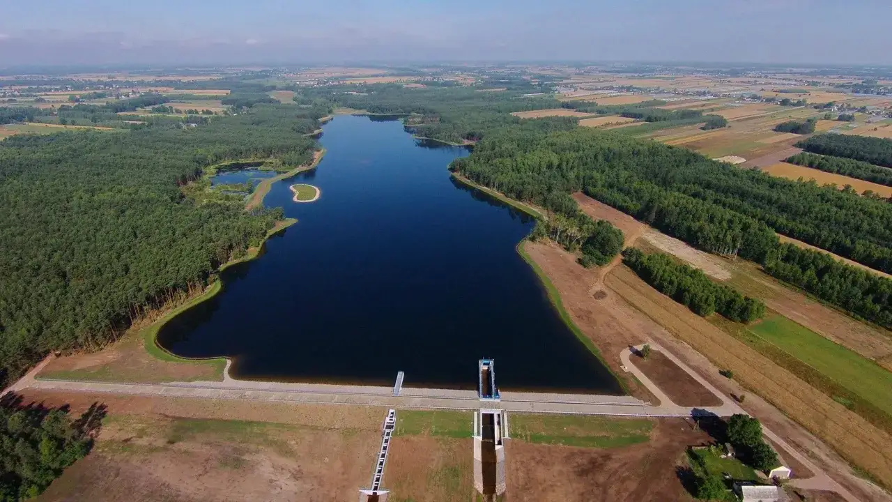 Widok z lotu ptaka na zalew Jagodno otoczony lasem i polami. Woda lśni, a tama z budowlami hydrotechnicznymi stanowi imponującą konstrukcję.