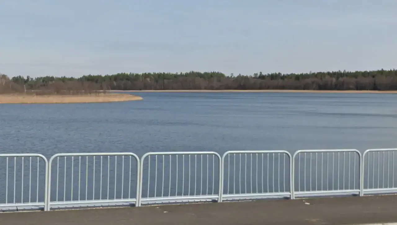 Spokojne jezioro Tyrkło otoczone lasem, z widoczną od strony promenady metalową balustradą.